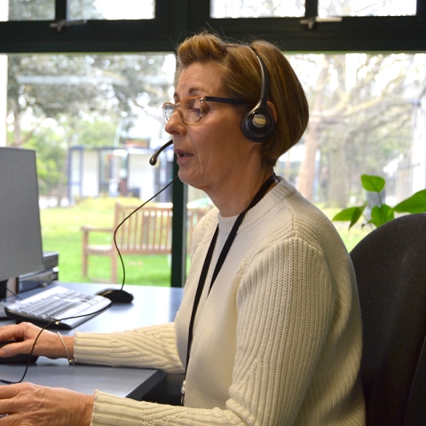 Female librarian sat at a computer in an office wearing a white cardigan and headset, answering a telephone call