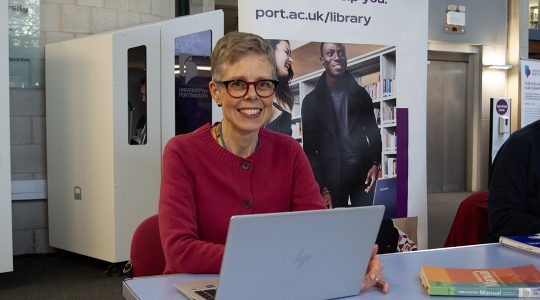CCI Faculty Librarian Greta Friggens sat in the Library Atrium in front of the Library pop-up banner