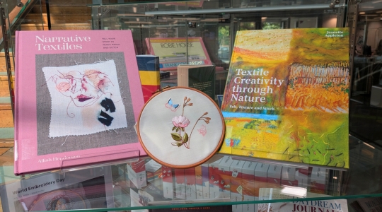 Embroidery depicting a butterfly visiting flowers sandwiched between library books about narrative embroidery and nature inspired textile design