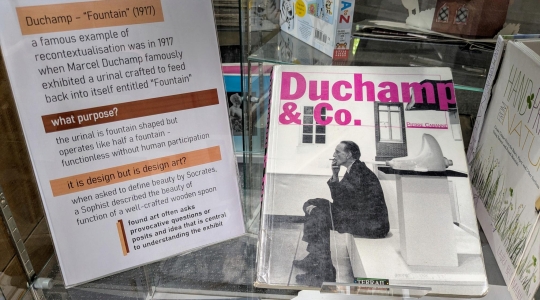Sign accompanied by a cover image of a book showing groundbreaking artist Marcel Duchamp sat smoking next to his 1917 urinal sculpture titled "Fountain"