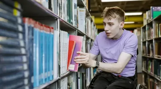 Young man in wheelchair picking a book from a Library bookshelf