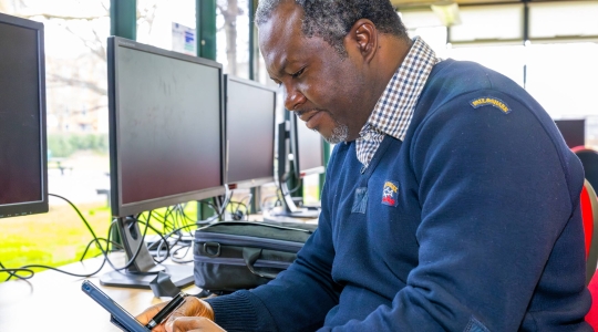 Black man sat at a computer in the Library Postgraduate Study Suite using a mobile phone
