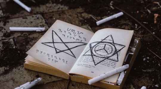 A ring of extinguished white wax candles surrounding an open book on which has been drawn arcane sigils and motifs - from Pexels, https://www.pexels.com/photo/opened-book-3050270/