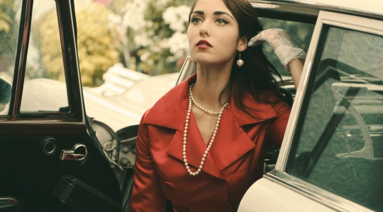 Woman in red coat and pearl necklace getting out of a vintage car, representing late 20th century womenswear.  From Pexels - https://www.pexels.com/photo/pretty-woman-posing-beside-a-vintage-car-8791108/ 