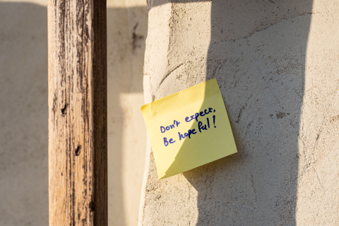 Post-it note attached to a pillar in dappled sunlight.  Note reads "Don't expect, be hopeful!".  From Pexels, https://www.pexels.com/photo/sticky-note-on-a-wall-in-sunlight-19664313/