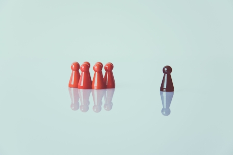 A cluster of red pawns opposite a lone Black pawn, all stood on a reflective surface.  From Pexels - https://www.pexels.com/photo/one-black-chess-piece-separated-from-red-pawn-chess-pieces-1679618/