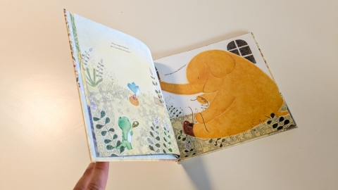A frog holding a leaf, a yellow elephant tying its shoelaces and a bluebird carrying a basket of berries are sown in a garden