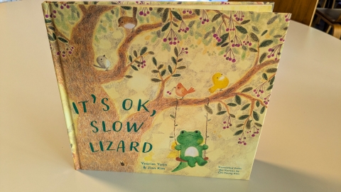 Cover of "It's ok, slow lizard" by Yeorim Yoon & Jian Kim, translated from the Korean by Chi-Young Kim.  Cover design shows a lizard swinging from a tree bearing berries and populated with birds.