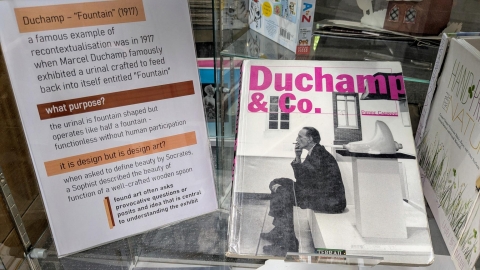 Sign accompanied by a cover image of a book showing groundbreaking artist Marcel Duchamp sat smoking next to his 1917 urinal sculpture titled "Fountain"