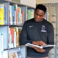 A student standing next to books in the library, reading an open book