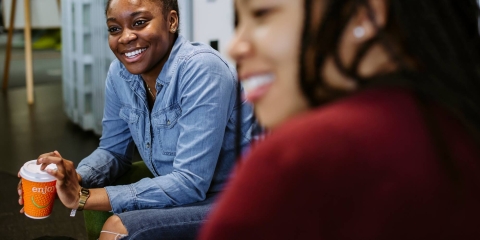 Two Black women sat in the Library Café, smiling and chatting with a third person outside of camera shot 