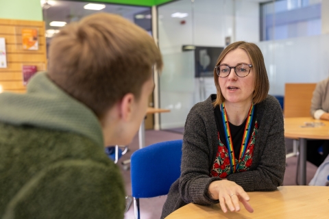 A staff member talking with a student at a table in a university learning space.